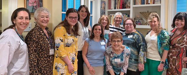 Eleven smiling women stand in a living room with a bookcase in the background